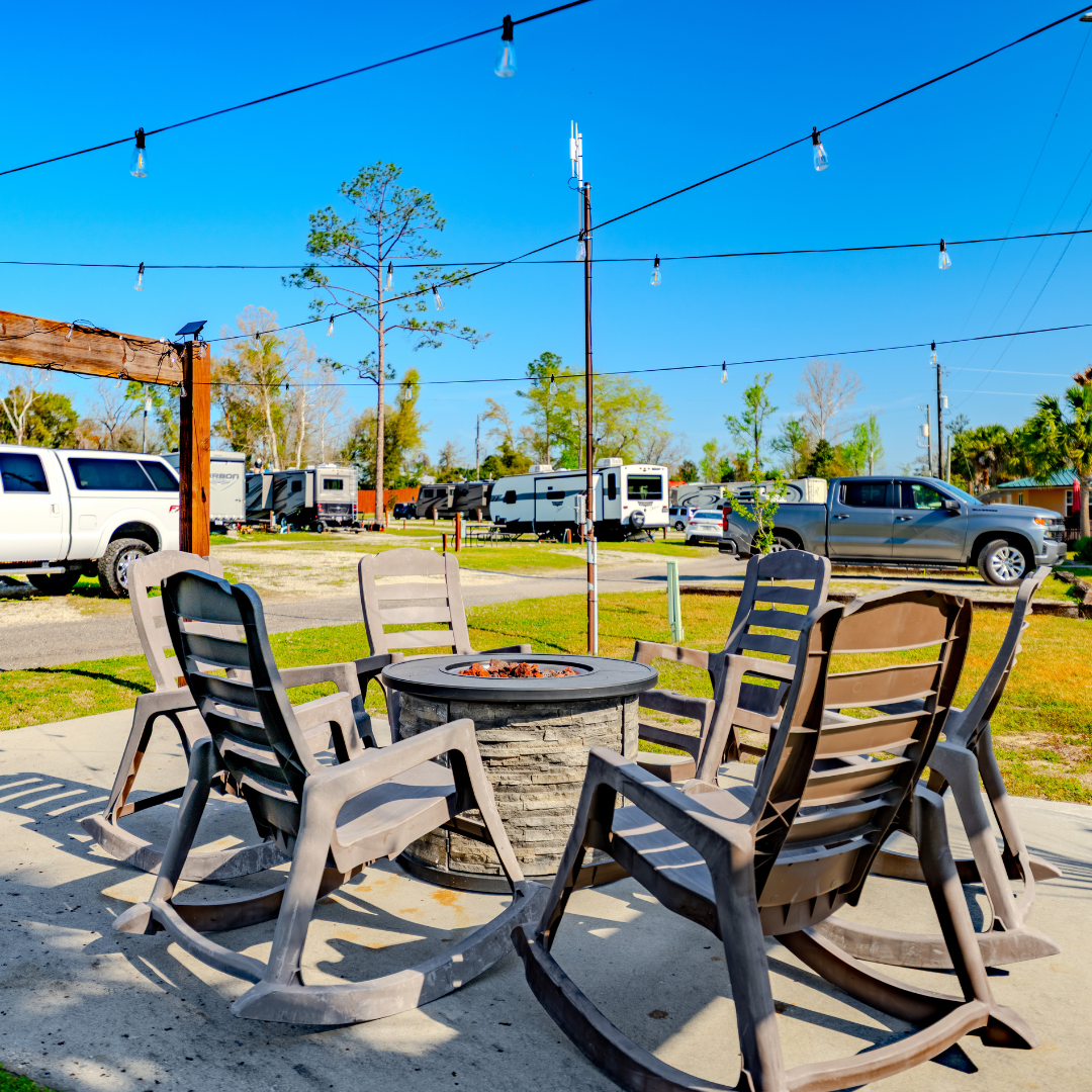 Photo of a circular outdoor fire pit with 6 chairs surrounding it. he sky in the back is bright blue, with same RVs and trucks parked on the grass.