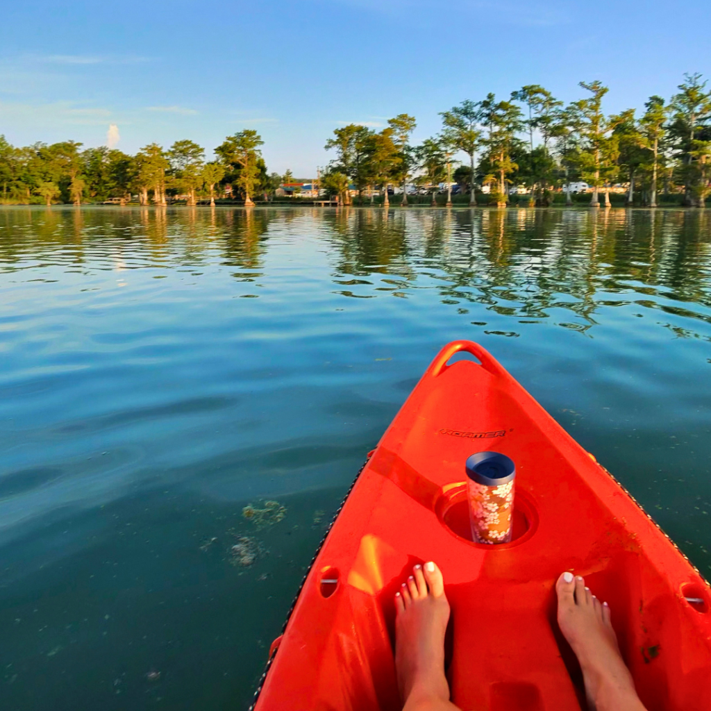 Photo of a kayak on the blue water, the front part of the kayak in only showing with feet sitting out on it and a cup in the small cup holder, trees in the background