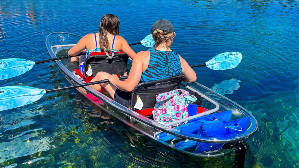 Photo of two women facing away from the camera kayaking in a clear kayak with a double-sided paddle in each of their hands. They are each looking to the right. The water is blue and crystal clear. They are on Merritt's Mill Pond with Get Up & Go Kayaking. This is in Marianna, Florida.
