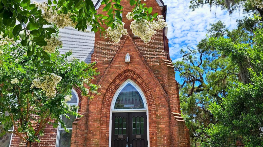 Photo of a church in Marianna, Florida with red brick and long doors with a window arch above. There are green leaves from a trees framing the photo. Cloudy blue skies in the background.